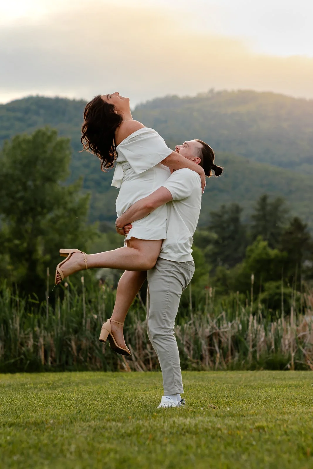 A man lifting a woman in a grassy field with mountains in the background, during sunset.