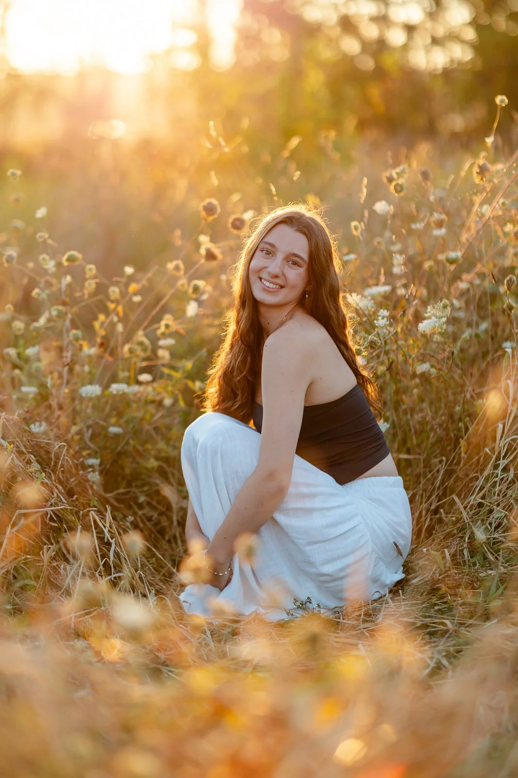 A young woman with long brown hair smiling while sitting in a field of wildflowers during sunset, wearing a black top and white pants.