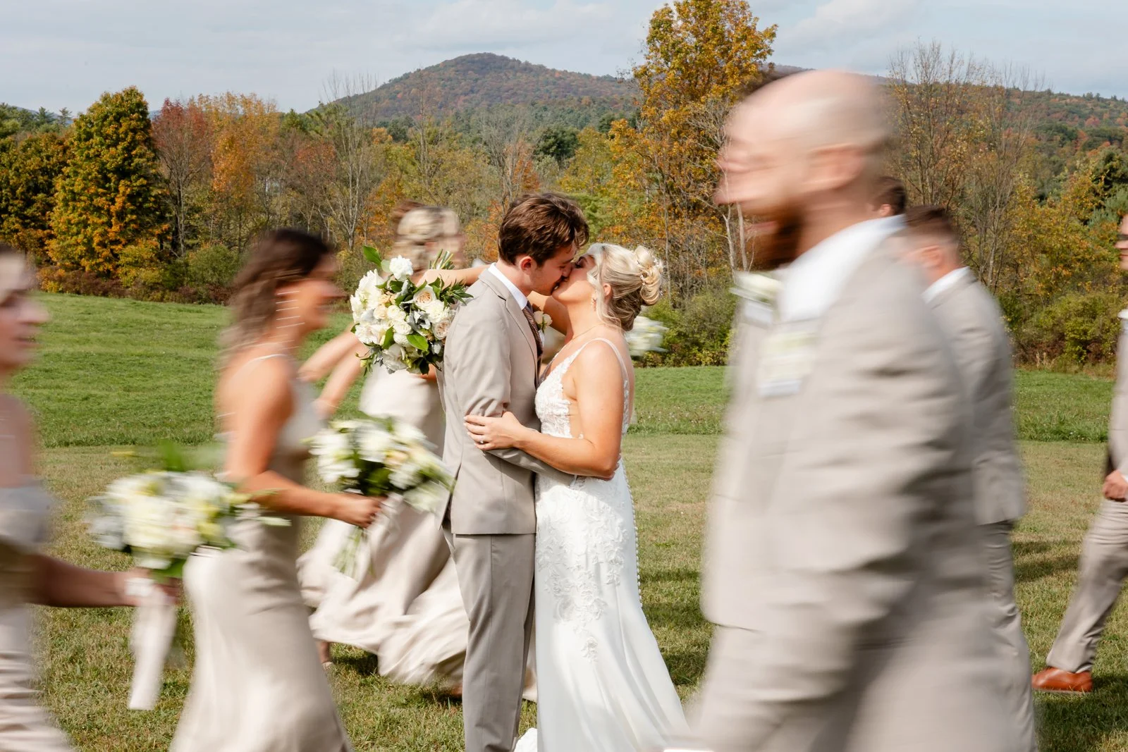 A newlywed couple kissing outdoors at their wedding, surrounded by bridesmaids and groomsmen, with colorful trees and mountains in the background.