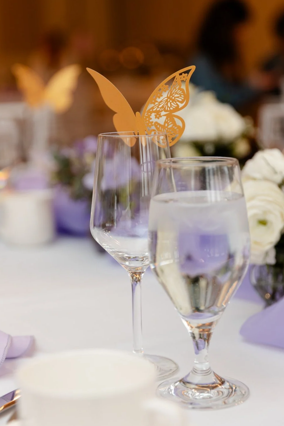 Elegant dinner table with two glasses of water, one decorated with a yellow butterfly paper decoration, surrounded by flowers and candle holders, suggesting a formal event or celebration.
