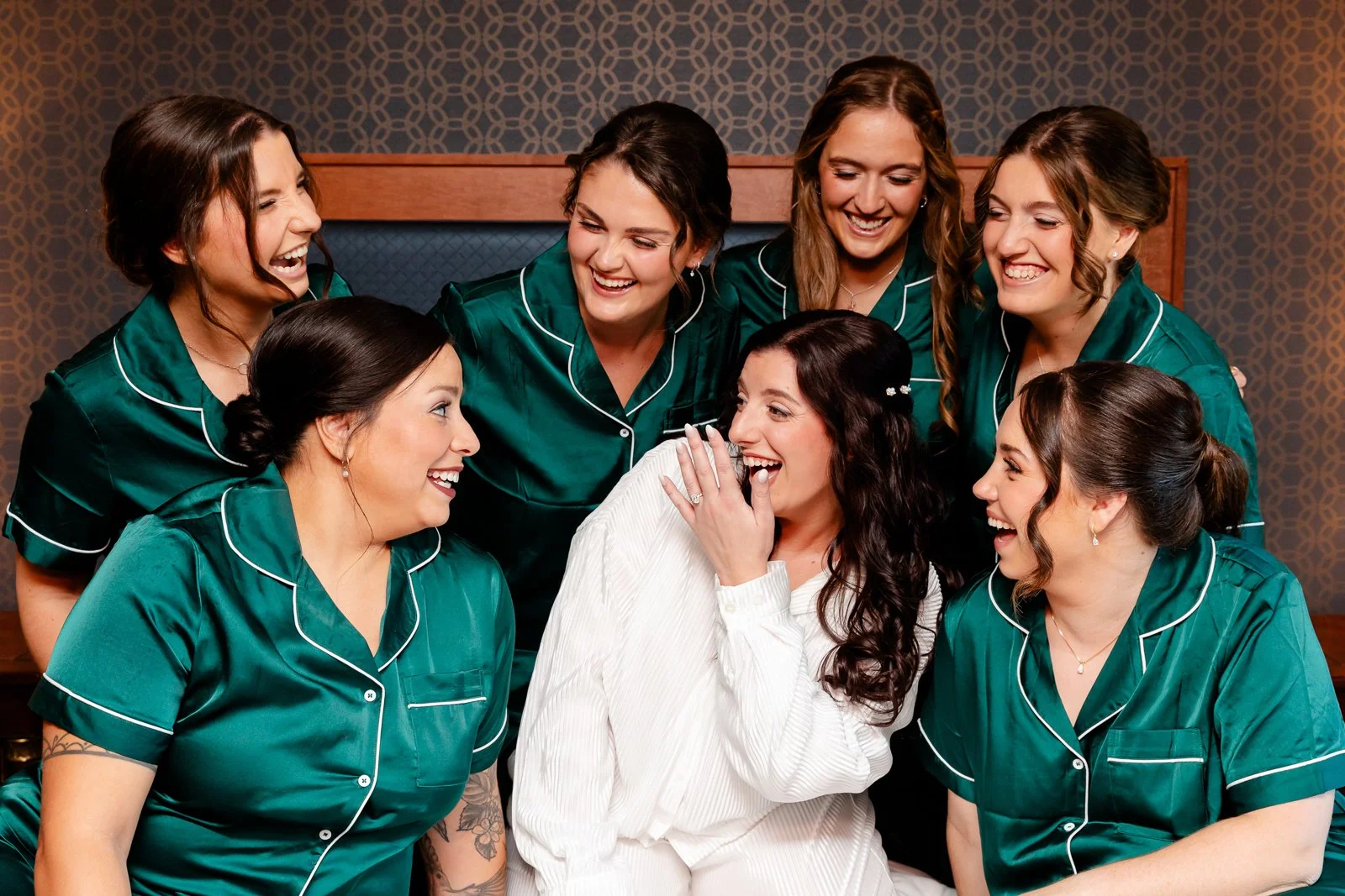 A bride in a white dress laughing and covering her mouth, surrounded by eight bridesmaids in matching green pajamas, all smiling and sharing a joyful moment.