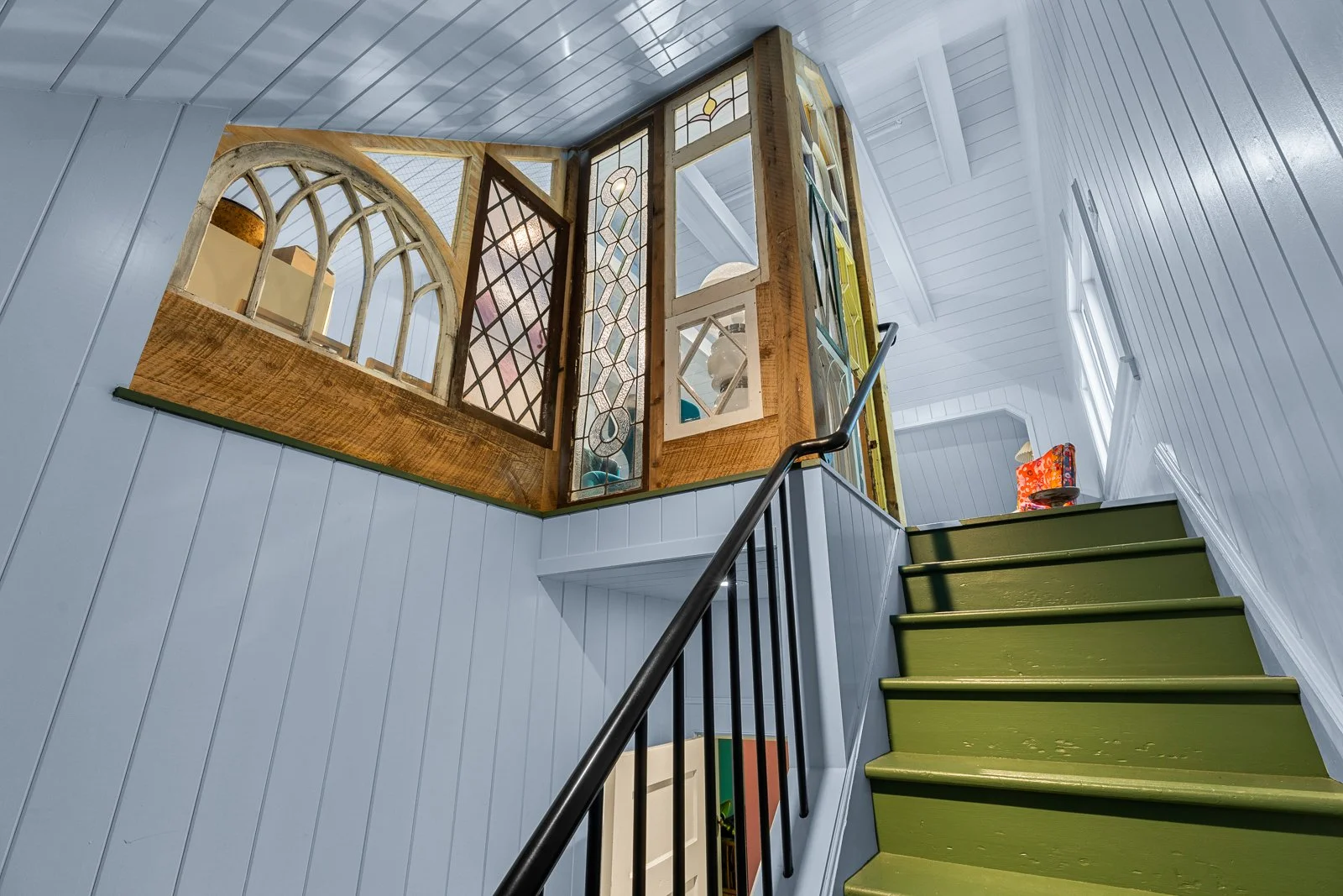 A staircase with green steps and a black handrail leading up to a landing with decorative stained glass windows and white wooden walls.