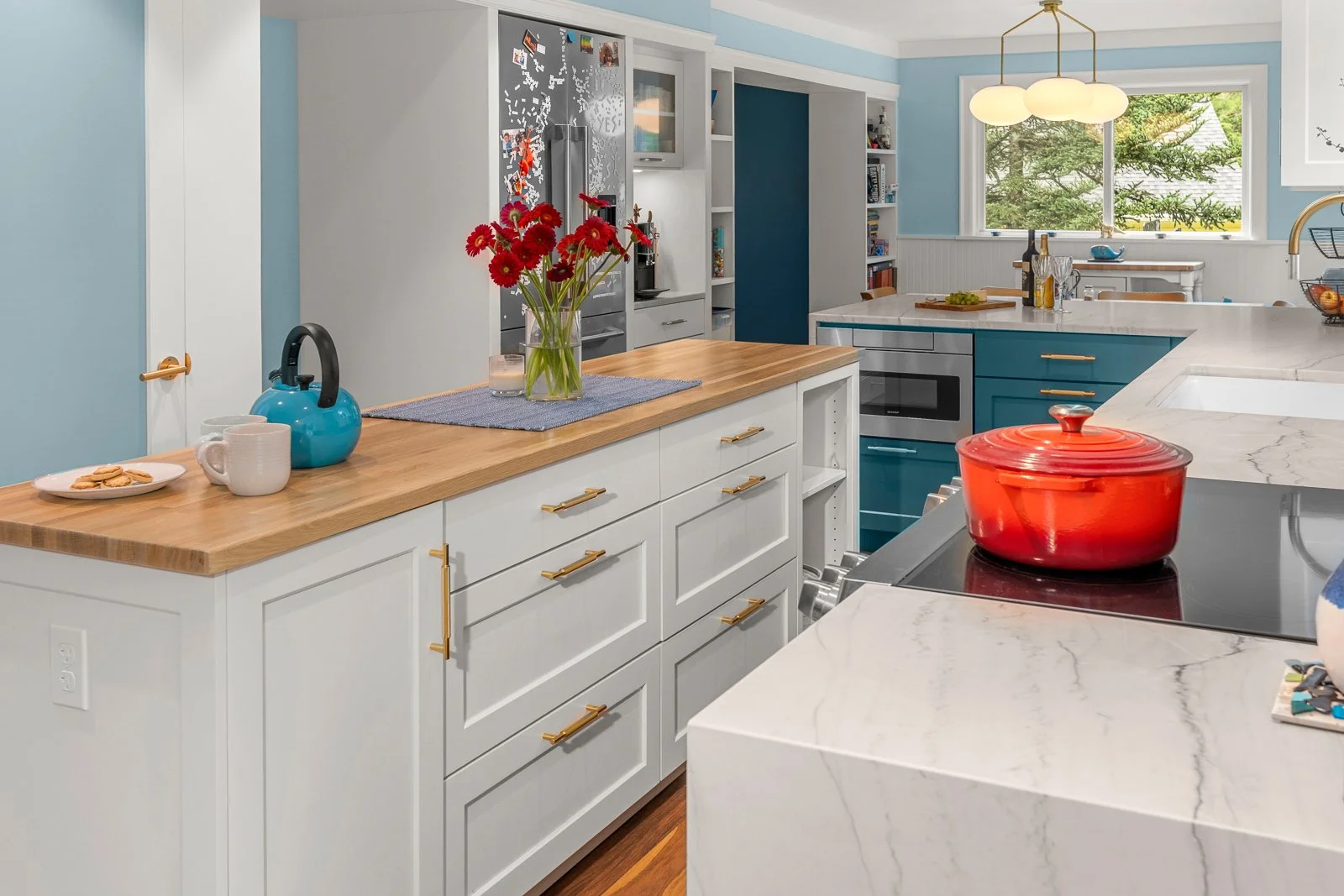 Modern kitchen with white and teal cabinets, a marble countertop, a red pot on the stove, a vase of red flowers on the island, and a window with greenery outside.