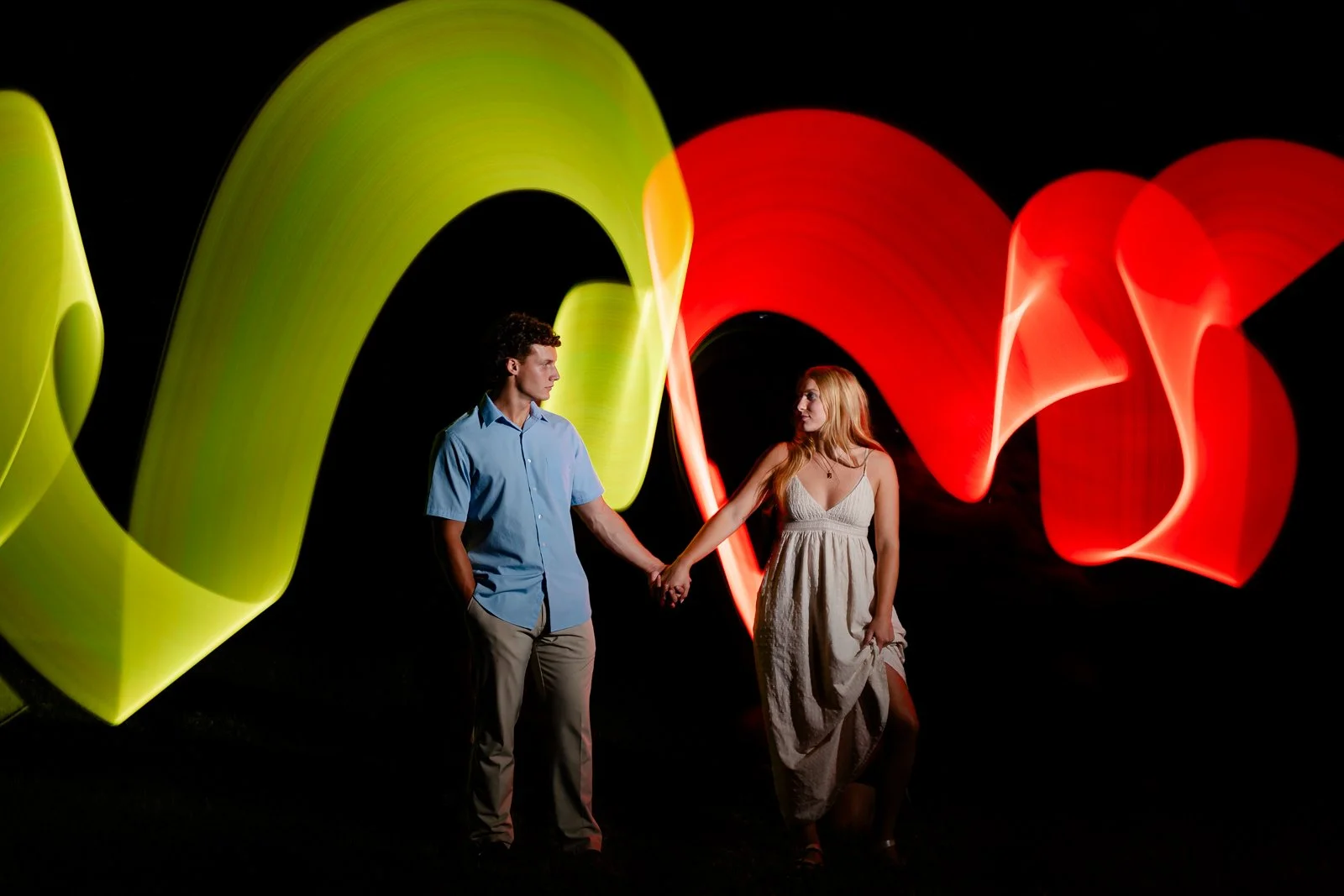A young man and woman holding hands in front of colorful light trails at night.