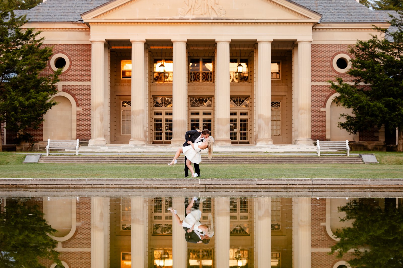 A couple is dancing in front of a large neoclassical building with tall columns, during sunset. Their reflection is visible in a pond in front of them.