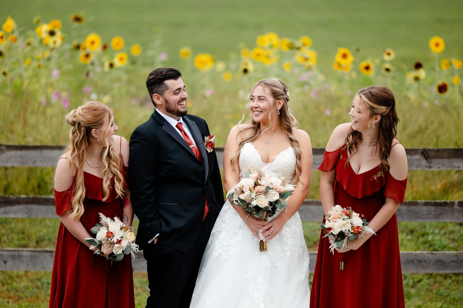 A wedding party with a bride, groom, and two bridesmaids standing outdoors in front of a wooden fence with a field of yellow flowers. The bride is wearing a white wedding dress and holding a bouquet, the groom is in a black suit with a red tie, and t
