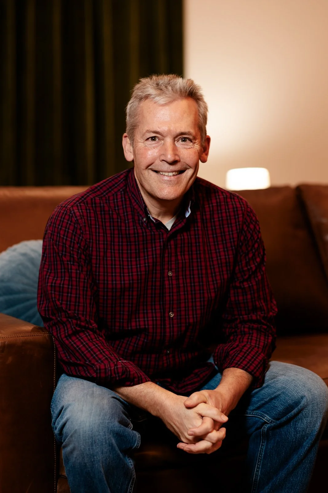 A smiling man with gray hair, wearing a red checkered shirt, sitting on a leather couch with a blue pillow in a warmly lit room.