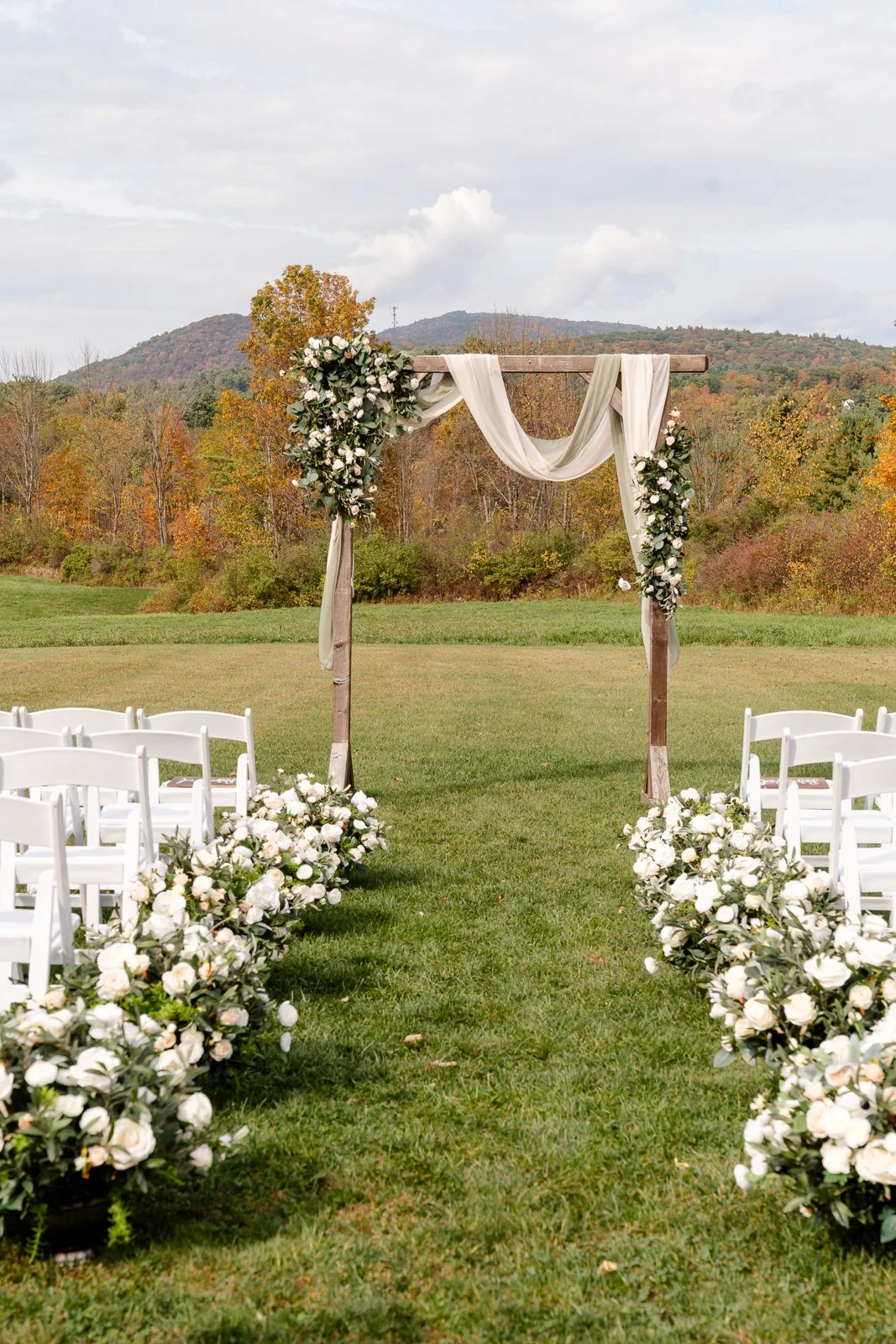 Outdoor wedding ceremony setup with a wooden arch decorated with white flowers and flowing fabric, surrounded by white chairs and floral arrangements, against a backdrop of trees and mountains.