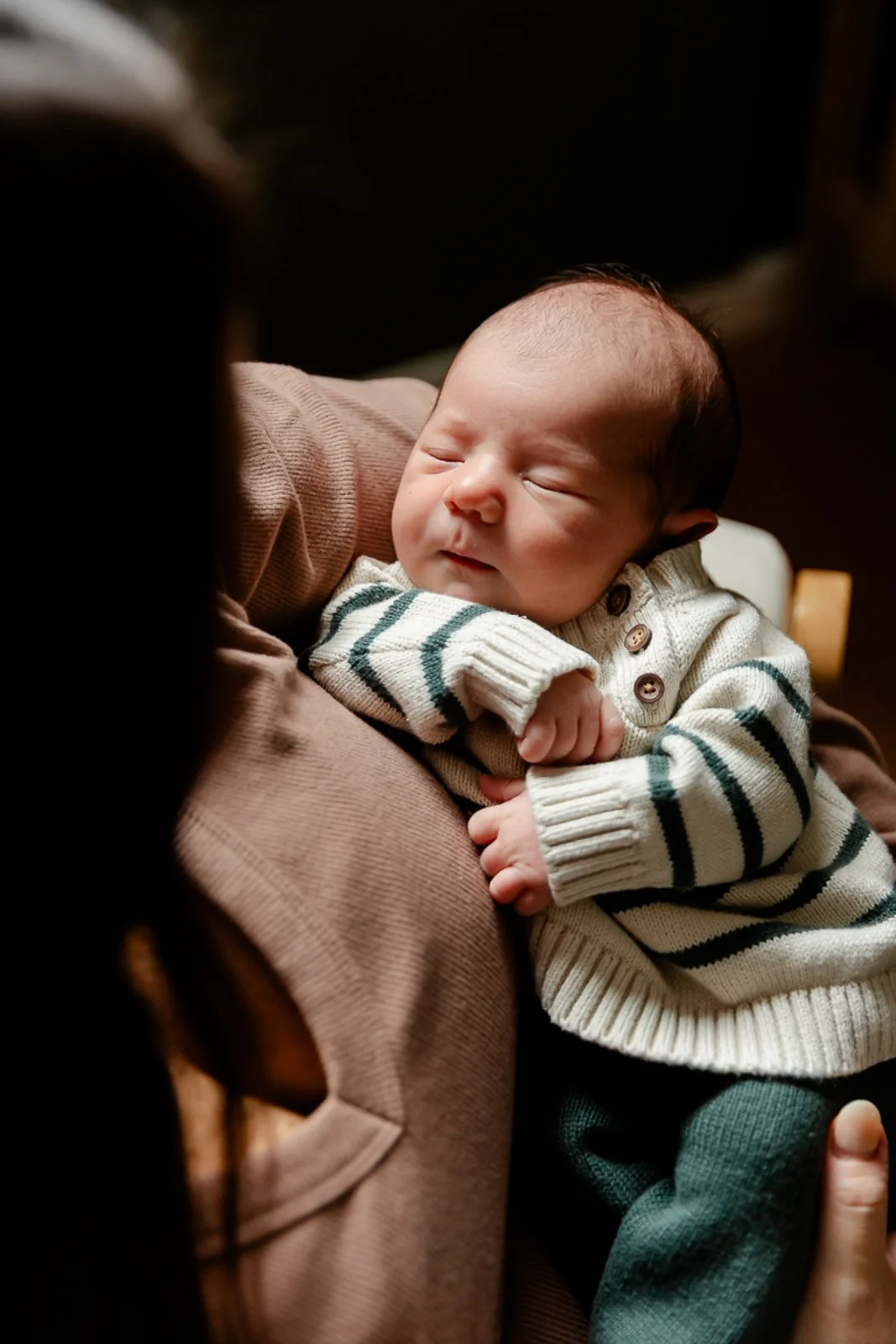 A sleeping baby held in an adult's arms, wearing a cream-colored sweater with black and green stripes and dark green pants.