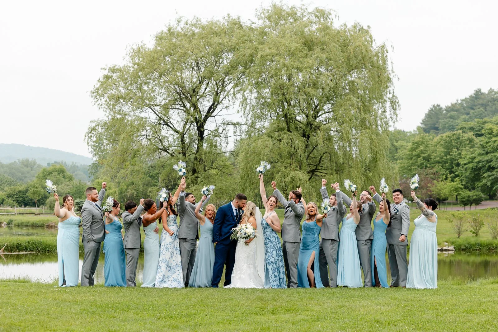 A wedding party standing outdoors near a pond with tall trees in the background, celebrating with raised arms and holding bouquets.