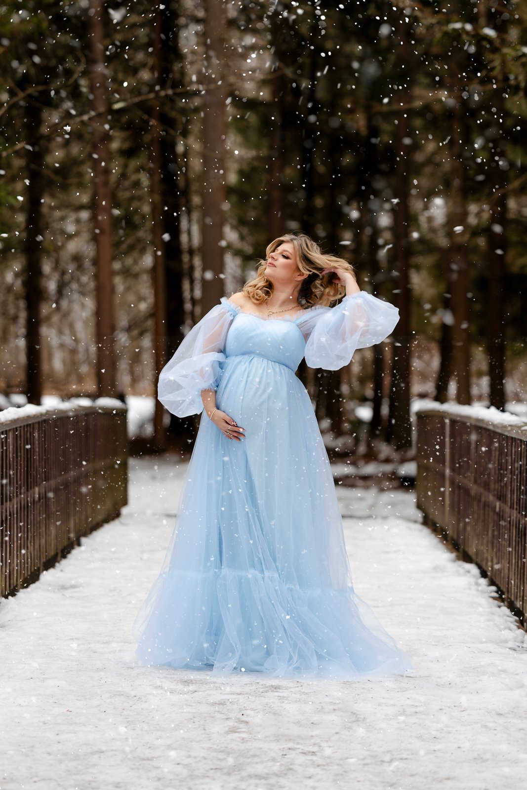 A woman in a long, light blue dress with puffy sheer sleeves standing on a snow-covered bridge surrounded by tall trees, snow falling around her.