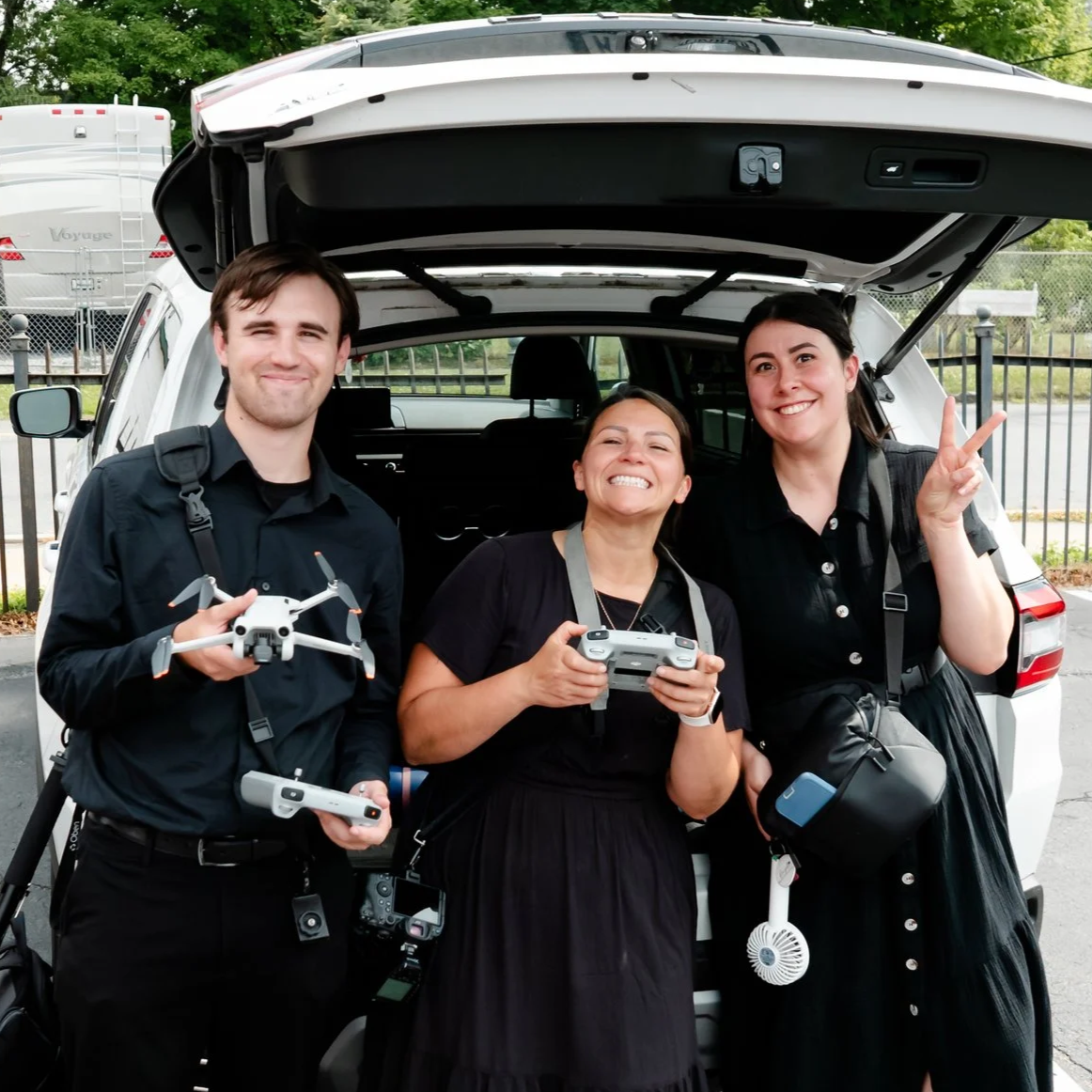 Three smiling women standing in front of an open car trunk, holding drone controllers and a drone, with a drone and camera strap around their necks, making peace signs and happy expressions.