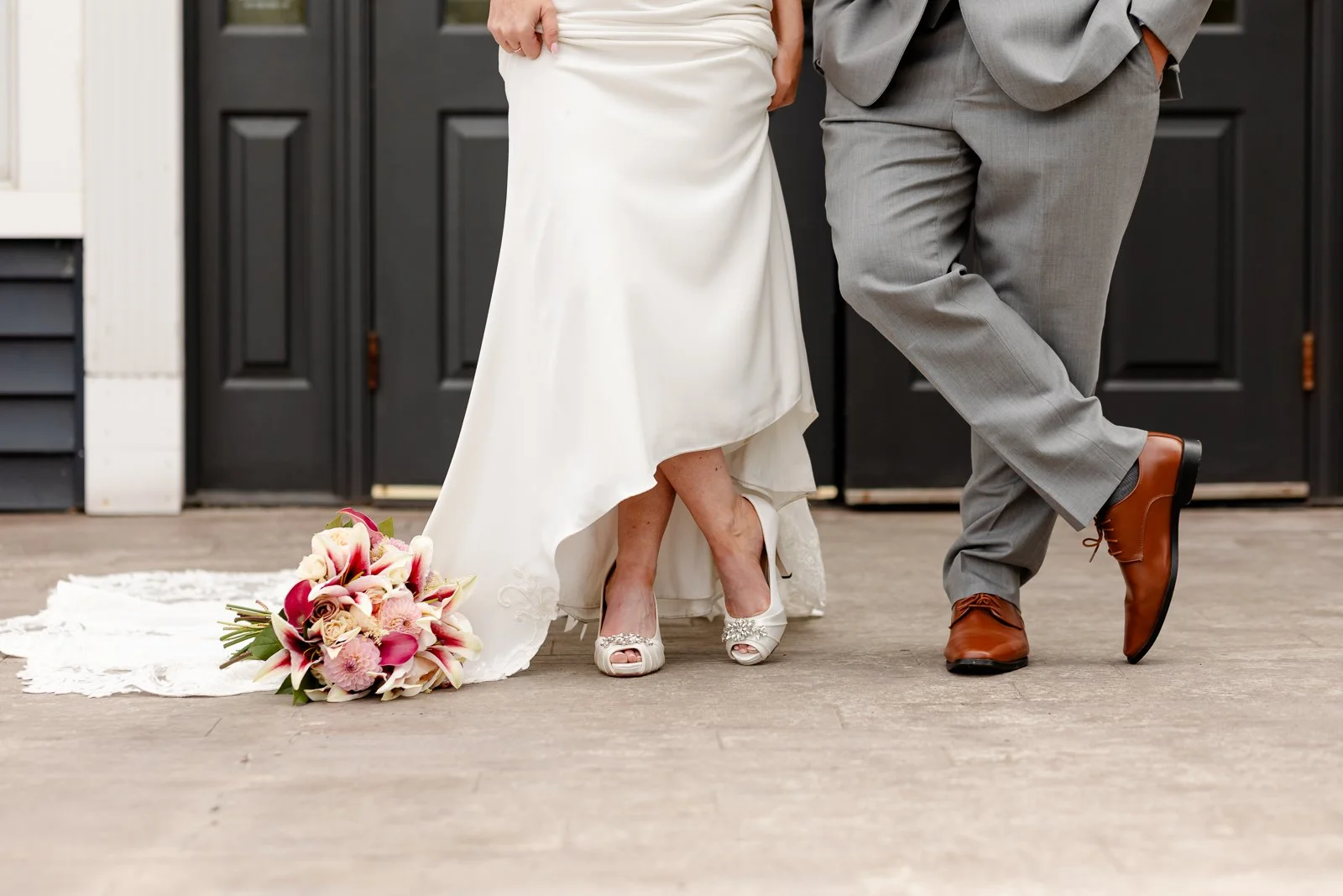 Bride and groom standing on a wooden floor, with the bride's bouquet on a white lace cloth, dress and shoes visible, and the groom in gray suit and brown shoes.