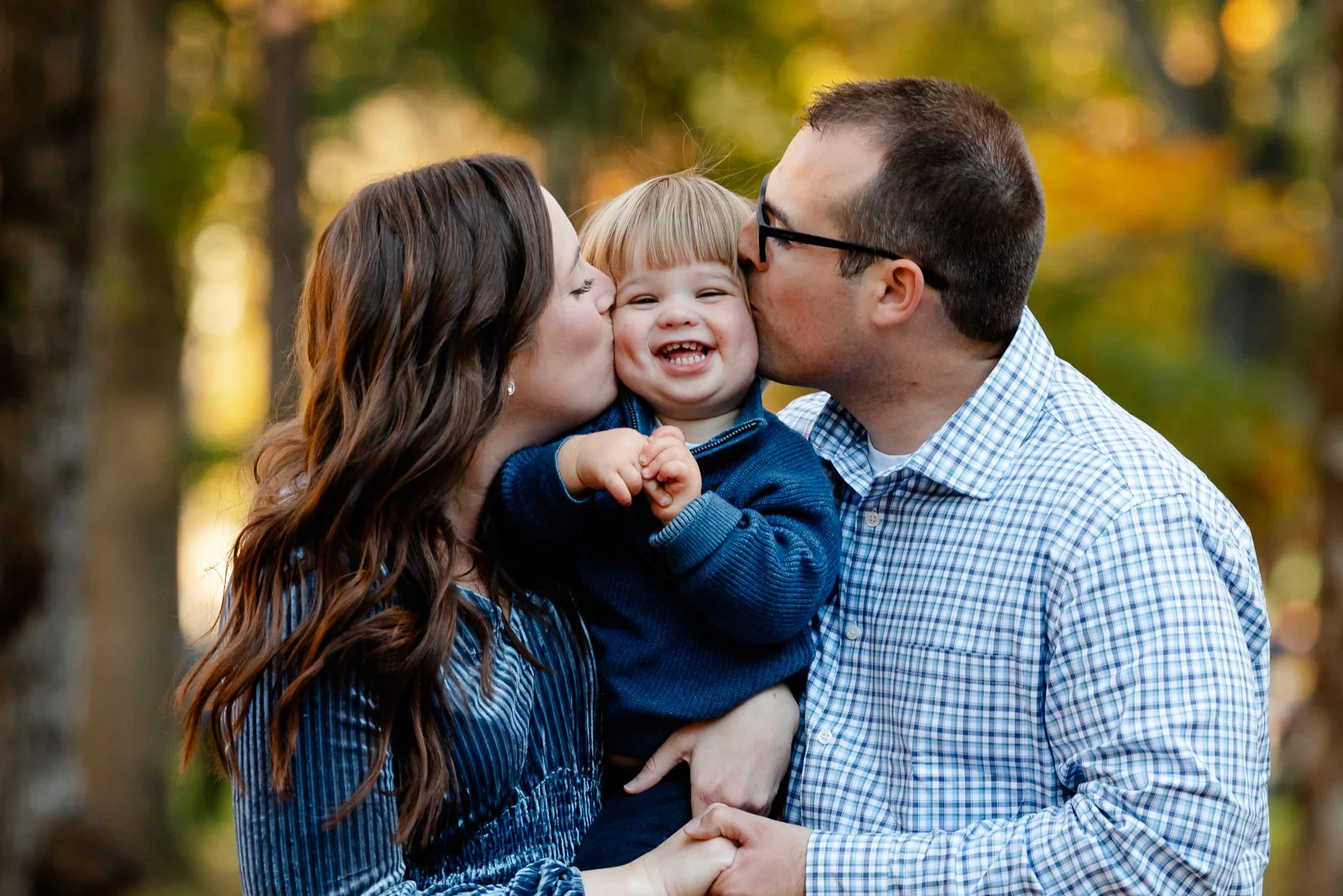 Family of three (mother, father, child) outdoors in a forest during autumn, with the parents kissing the child's cheeks, who is smiling and holding their hands.