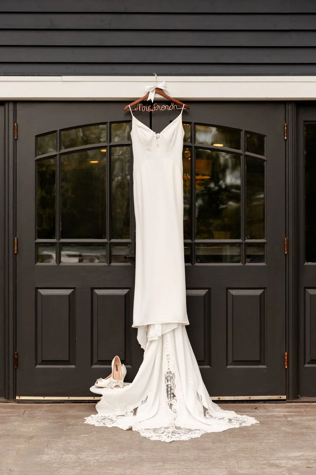 White wedding dress hanging on a hanger with a white ribbon on a black door, with white heels placed on the ground below, and a personalized hanger with the name 'Mrs. Frendh'.