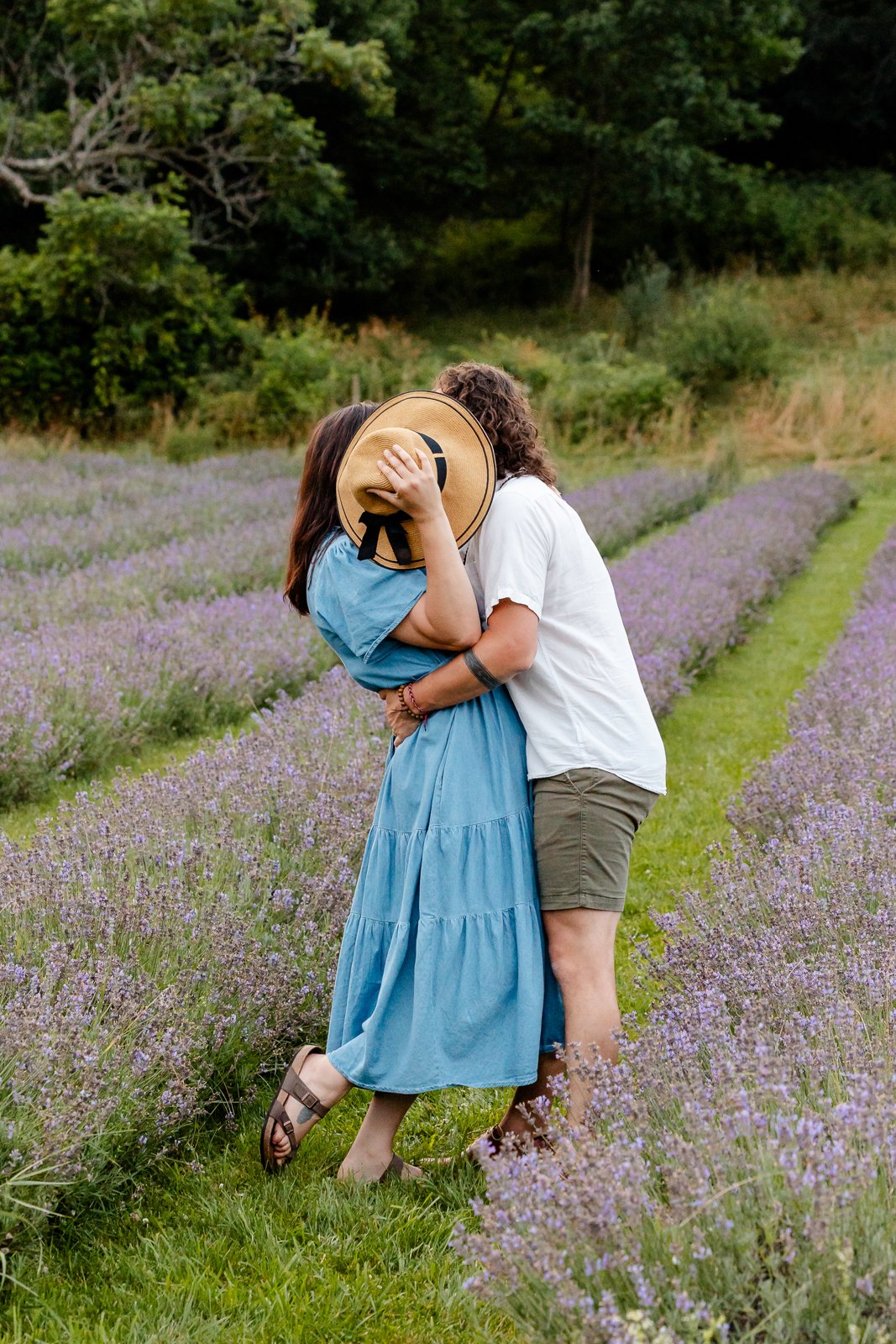 A couple kissing in a lavender field, with the woman holding a straw hat over both of their faces.
