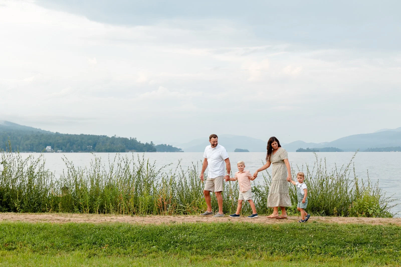 A family of five walking along a lakeside trail, holding hands, with water, mountains, and cloudy sky in the background.