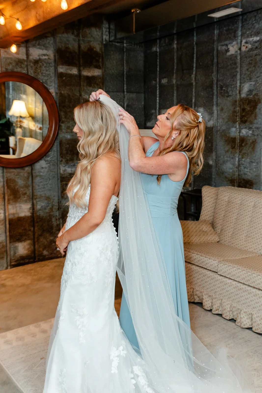 A woman in a white wedding dress is being assisted with her veil by another woman in a light blue dress in a cozy, rustic room with dark stone walls and a sofa.
