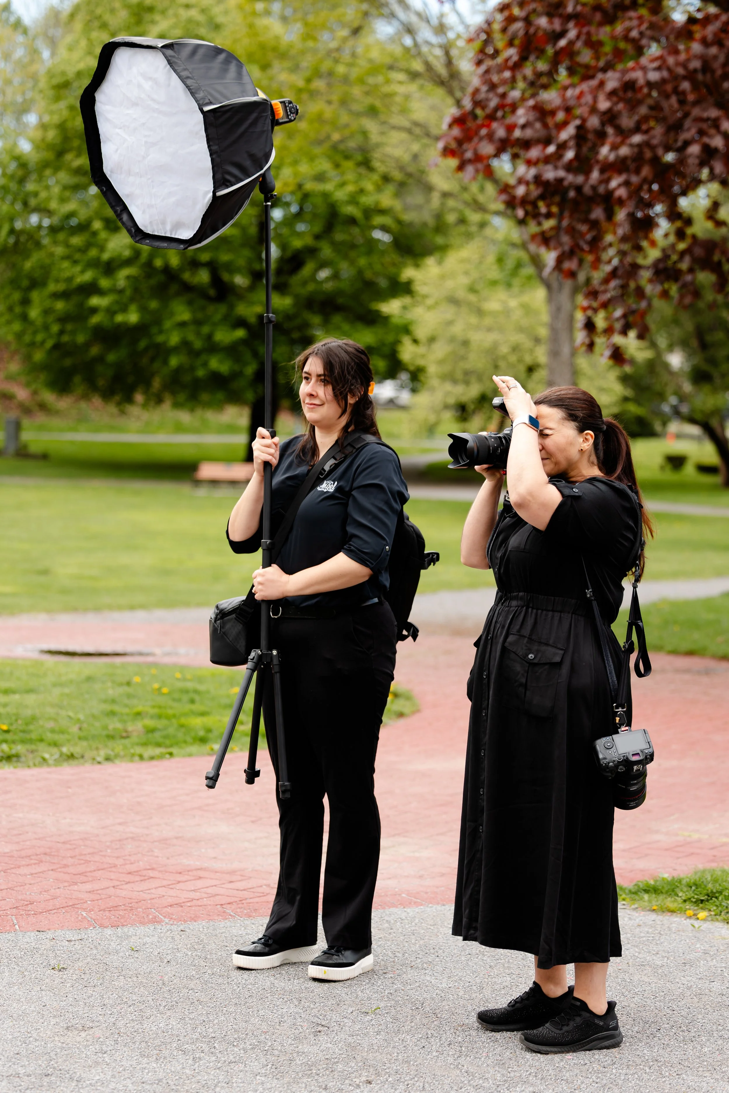 Two women, one holding a photography umbrella and a tripod, the other taking pictures with a camera, standing outdoors in a park with green trees around.