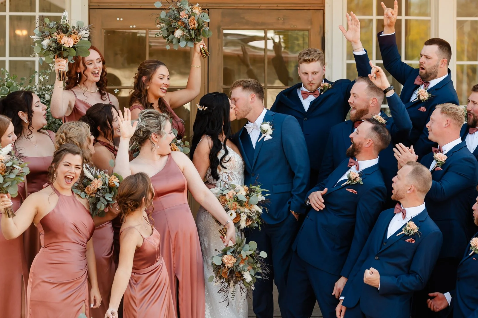A wedding celebration with the bride and groom sharing a kiss surrounded by their wedding party, including bridesmaids in mauve dresses and groomsmen in navy suits, some raising their hands in celebration.