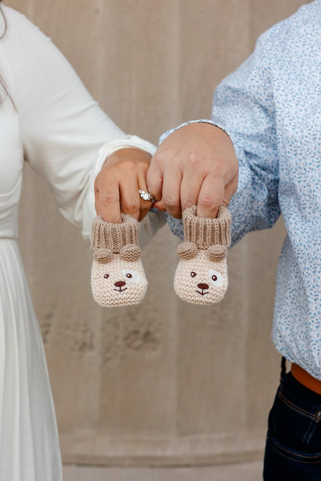Close-up of two hands holding knitted baby mittens shaped like bears, with one person wearing a wedding ring and beige sweater, and the other in a fitted white dress and a blue patterned shirt, against a neutral brown background.