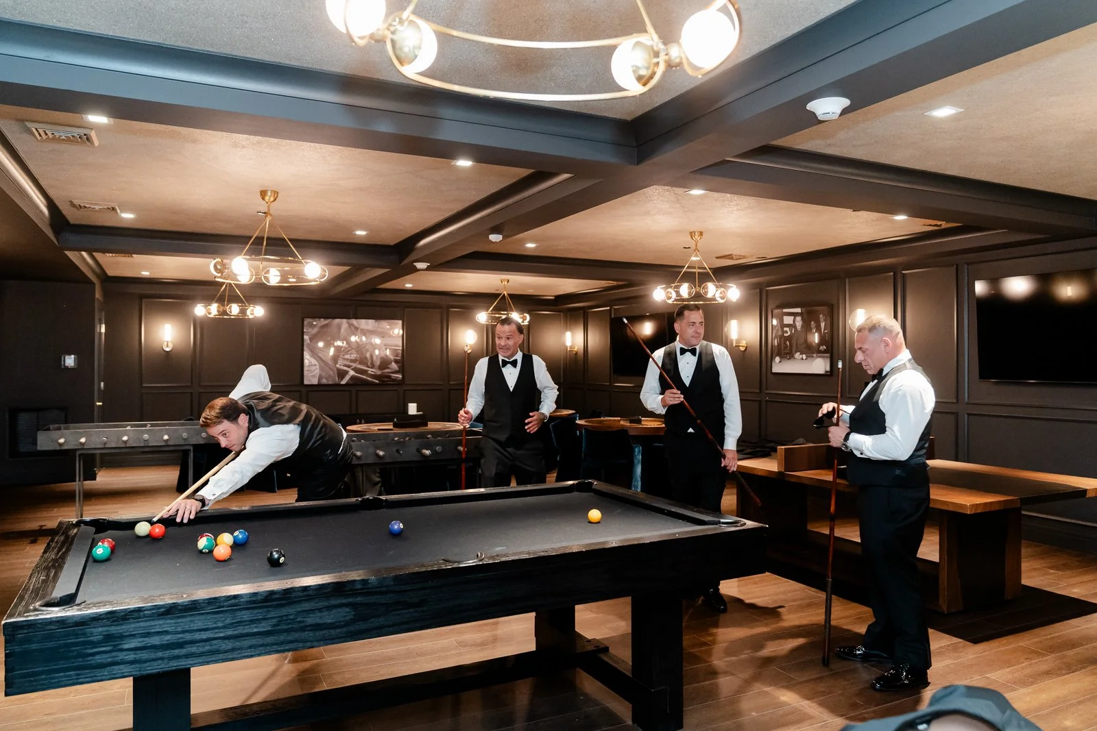 Four men in tuxedos and vests playing billiards in a dimly lit game room with dark walls and modern lighting fixtures.
