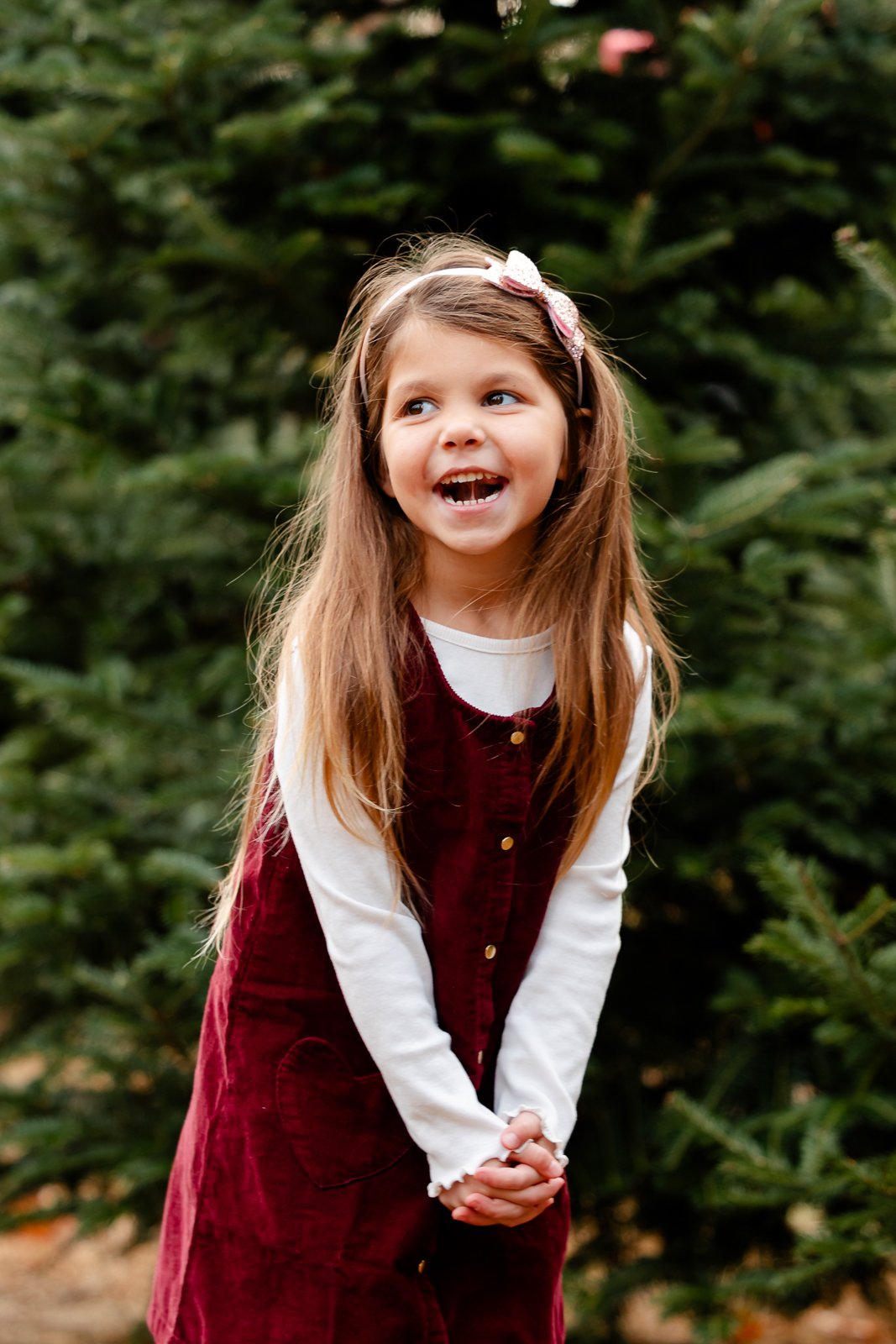 A young girl with long brown hair, wearing a white long-sleeve shirt and a maroon dress, smiling and clasping her hands together outdoors with green trees in the background.