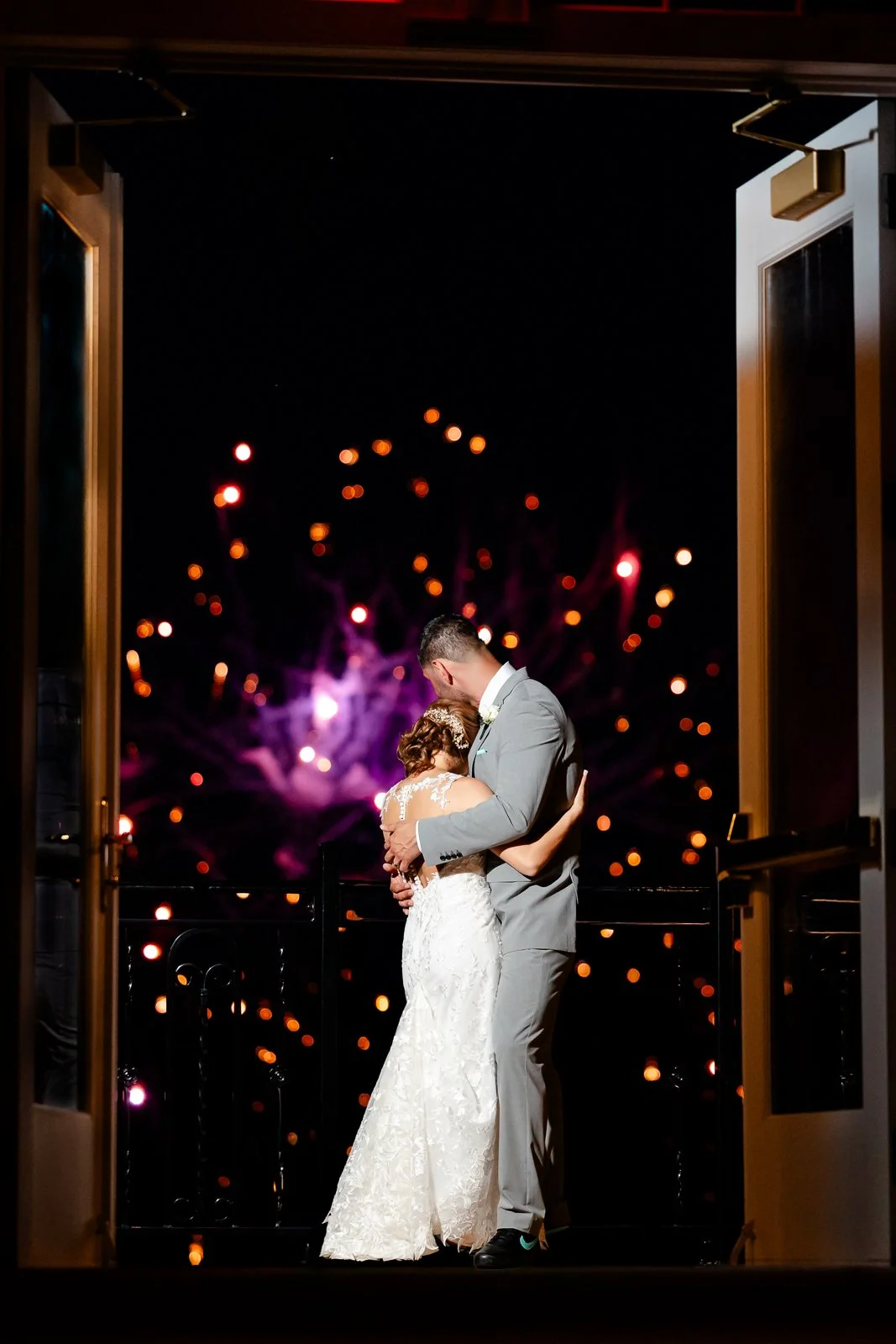 A bride and groom embrace on a balcony during fireworks at night.