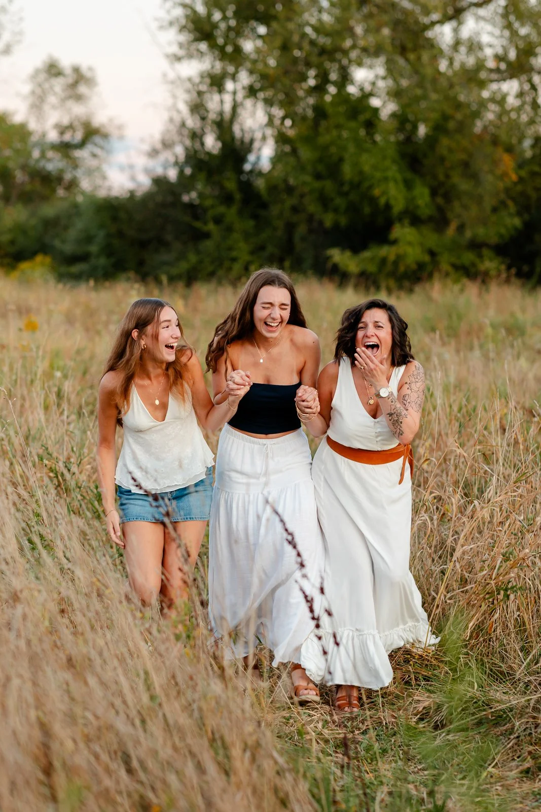 Three women walking and laughing in a field of tall grass during daytime, surrounded by trees.