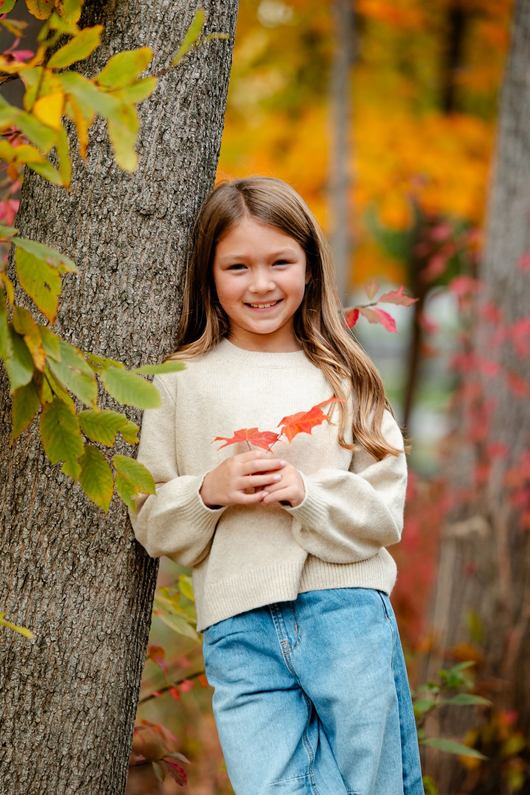 A young girl with long brown hair, wearing a cream sweater and blue jeans, stands outdoors leaning against a tree. She is holding autumn leaves and smiling at the camera, with a background of colorful fall foliage.