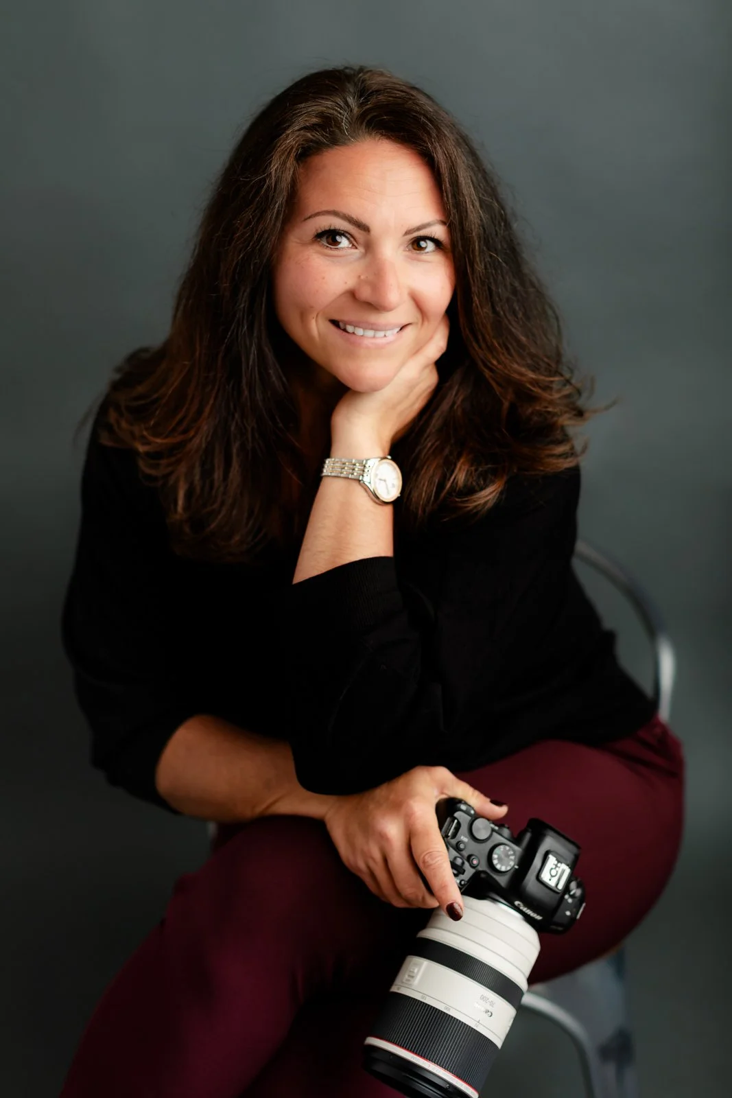 A woman with long brown hair and a watch on her left wrist, smiling and resting her chin on her hand, holding a professional camera with a large lens in her right hand, seated on a gray chair against a dark gray background.