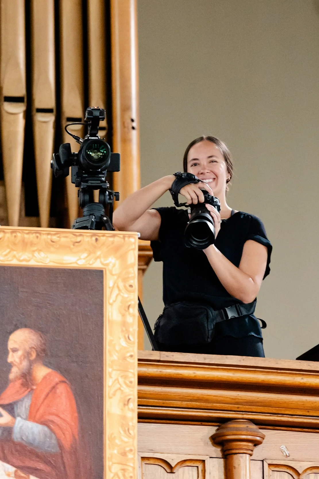 A woman smiling and holding a camera, standing next to a professional video camera on a tripod in an indoor setting with wooden architectural details.