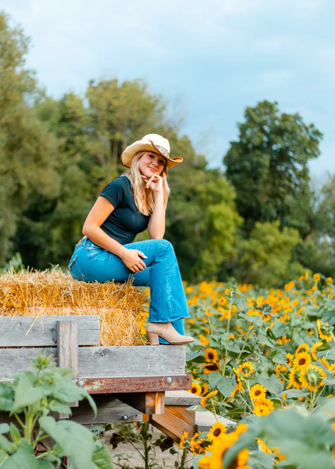 A woman sitting on hay bales on a farm surrounded by yellow sunflowers, wearing a cowboy hat, black shirt, blue jeans, and beige boots.