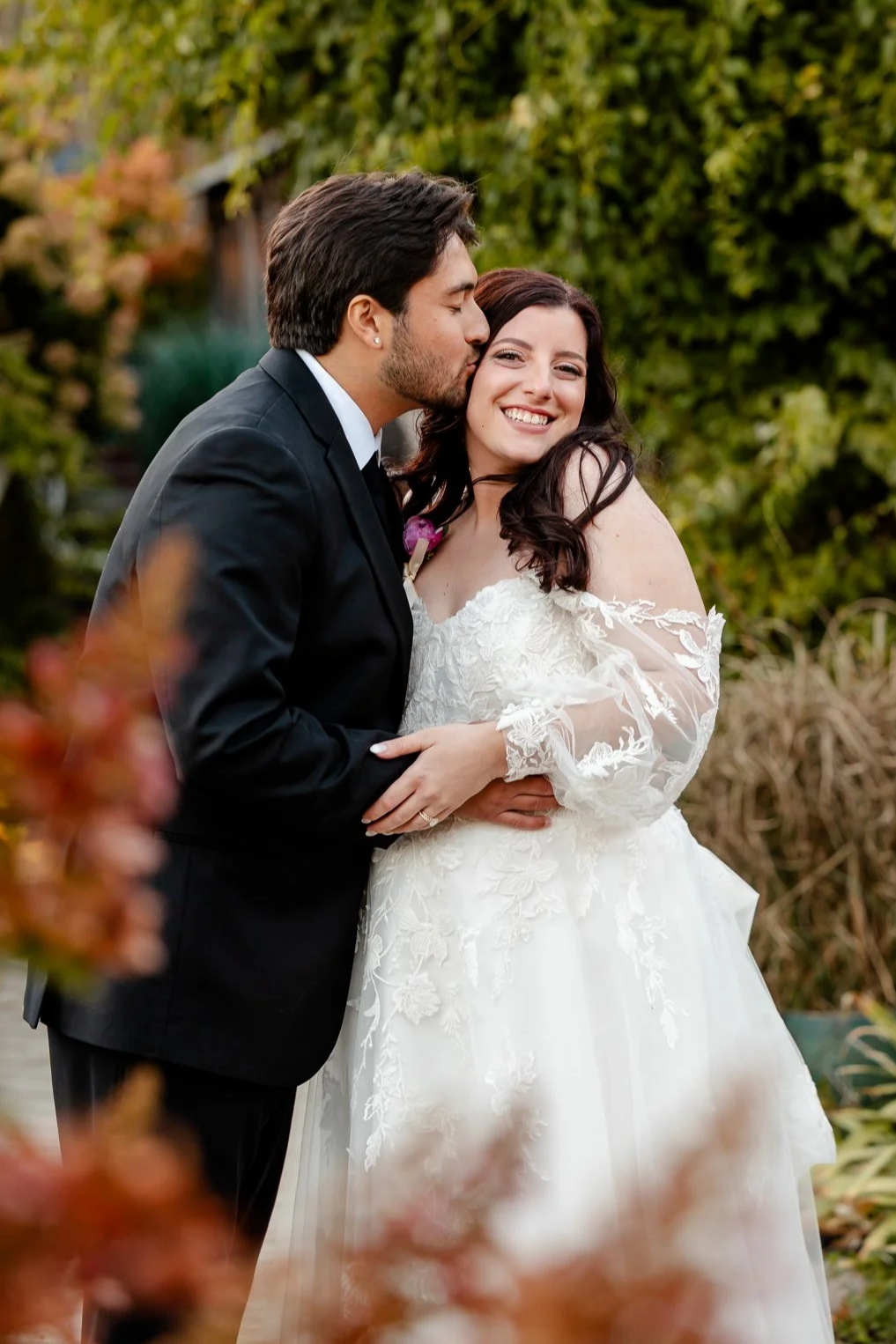 A bride and groom sharing a kiss outdoors, with the groom kissing the bride on the forehead, and the bride smiling, surrounded by greenery.