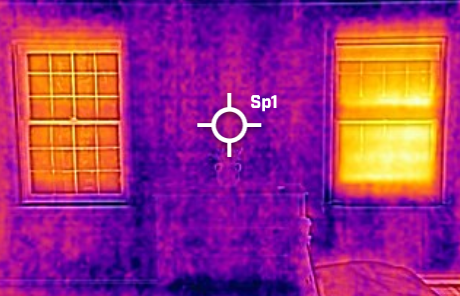 Infrared thermal image of a house wall with two windows emitting heat; the wall and frames appear in purple and yellow indicating temperature differences.