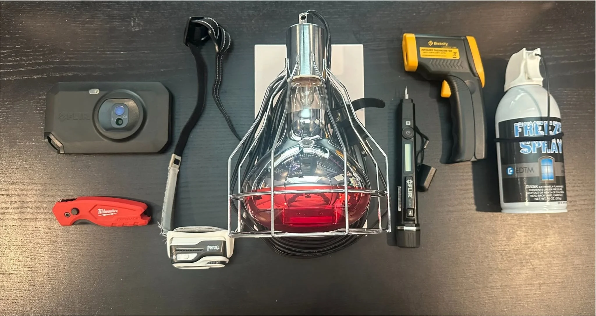 Collection of tools and equipment on a dark wooden surface, including a battery pack, a headlamp, a thermal scanner, a spray bottle labeled 'Freeze Spray', a utility knife, a lanyard, and a small electronic device with a screen.