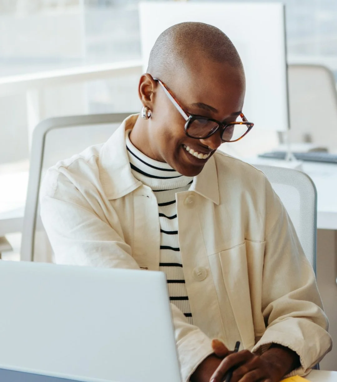A smiling woman with a short buzz cut hairstyle, wearing glasses, a striped turtleneck, and a light-colored jacket, sitting at a desk in an office.