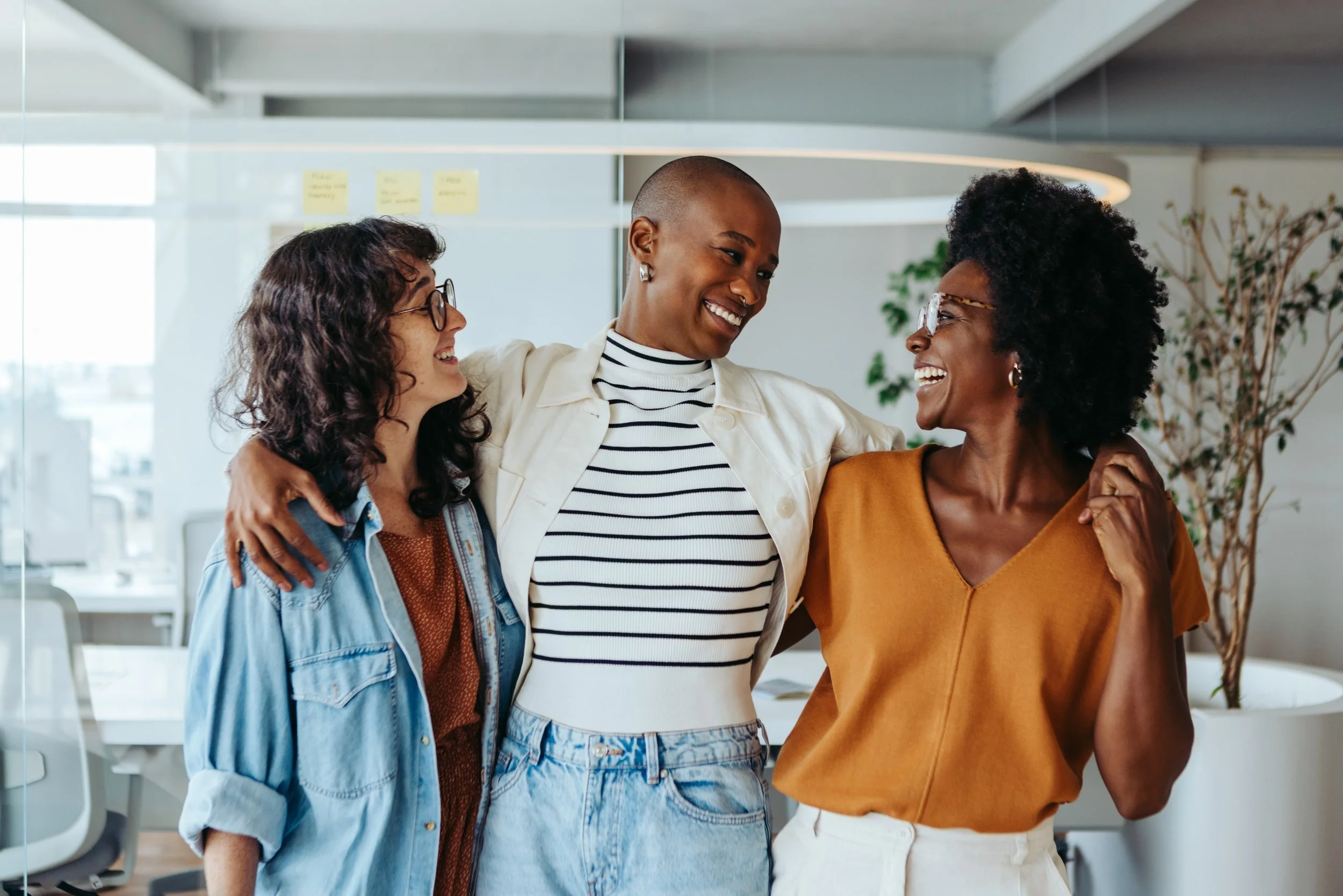 Three women are smiling and hugging each other in an office environment.