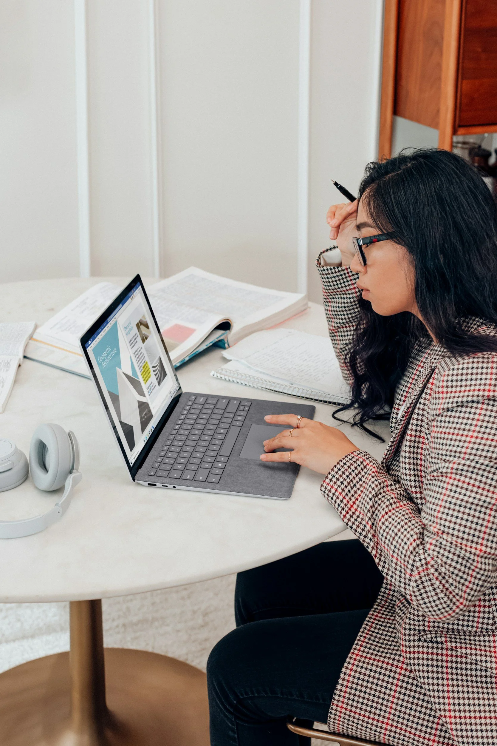 A woman with dark hair and glasses sitting at a white table, working on a laptop with multiple open books and notebooks around her.
