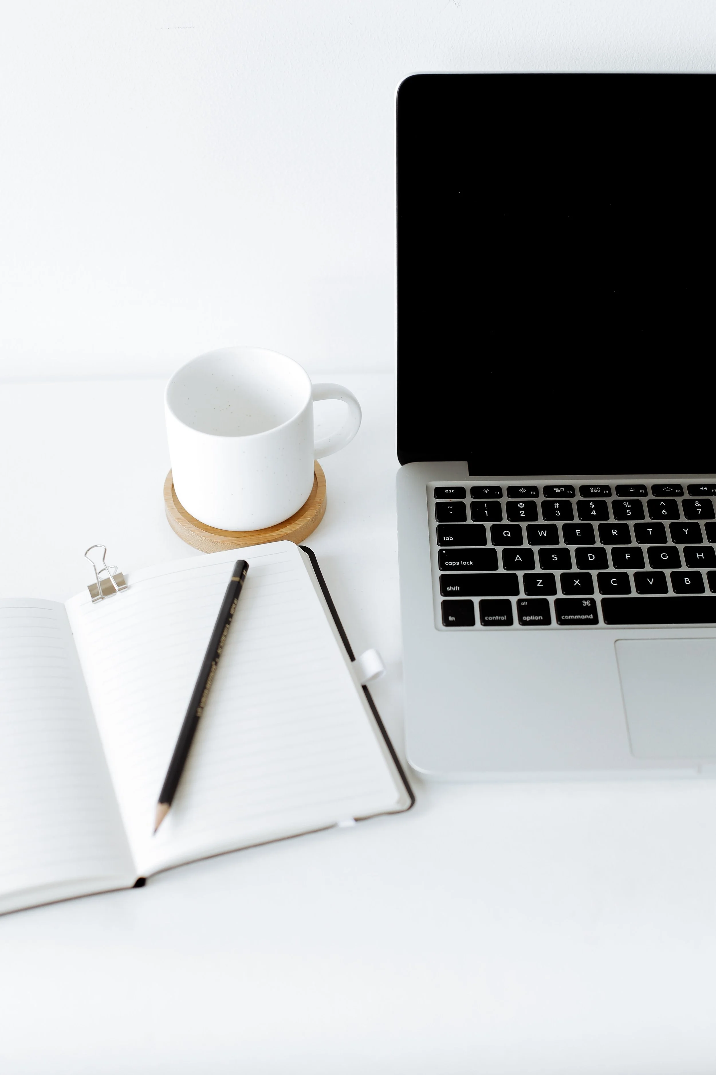 Work desk with a laptop, open notebook with a pencil, a white coffee mug on a wooden coaster, and a binder clip on the notebook, against a plain white background.