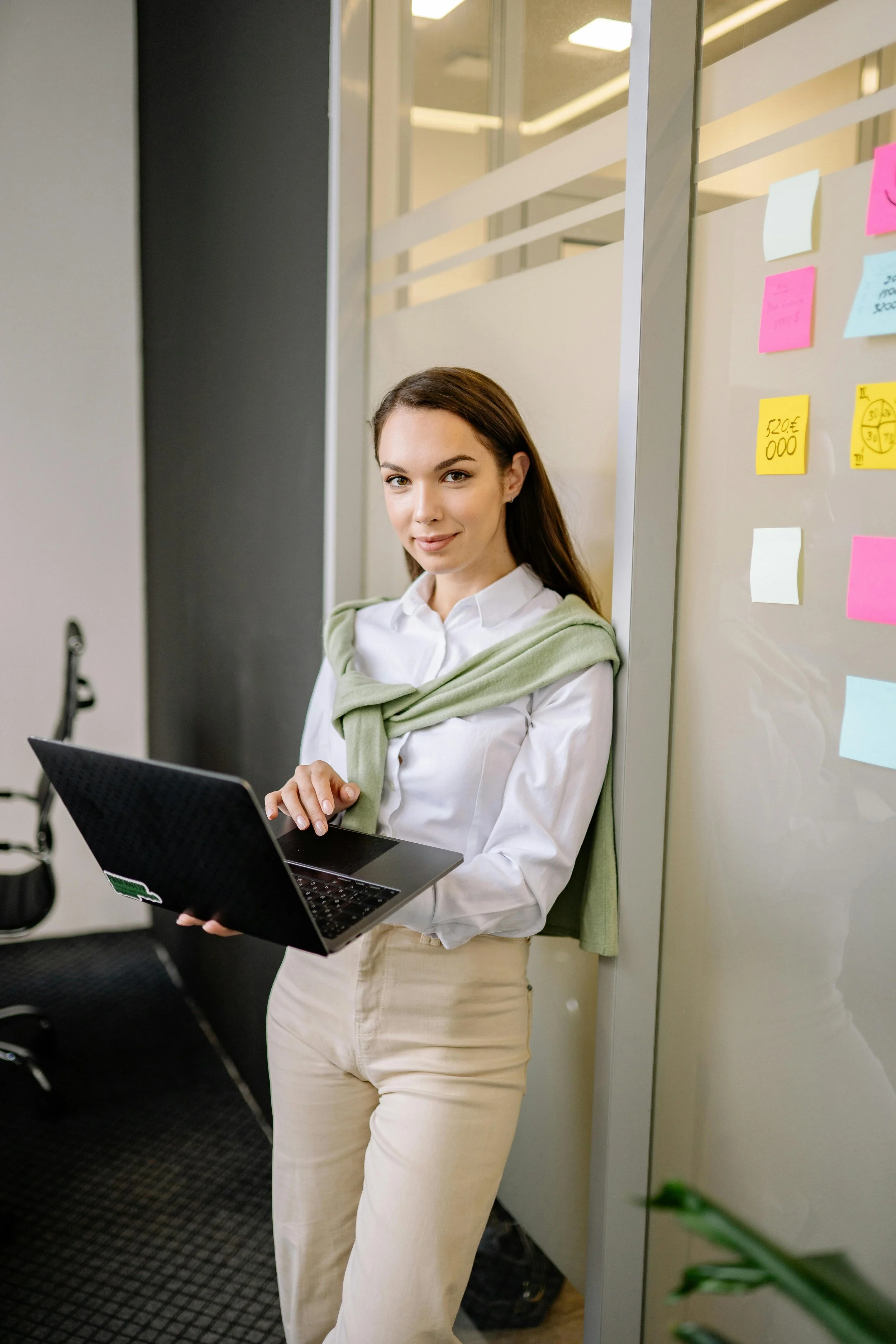 A woman with brown hair, wearing a white blouse and beige pants, standing in an office, holding a laptop. She has a light green sweater draped over her shoulders and stands near a glass wall with colorful sticky notes.