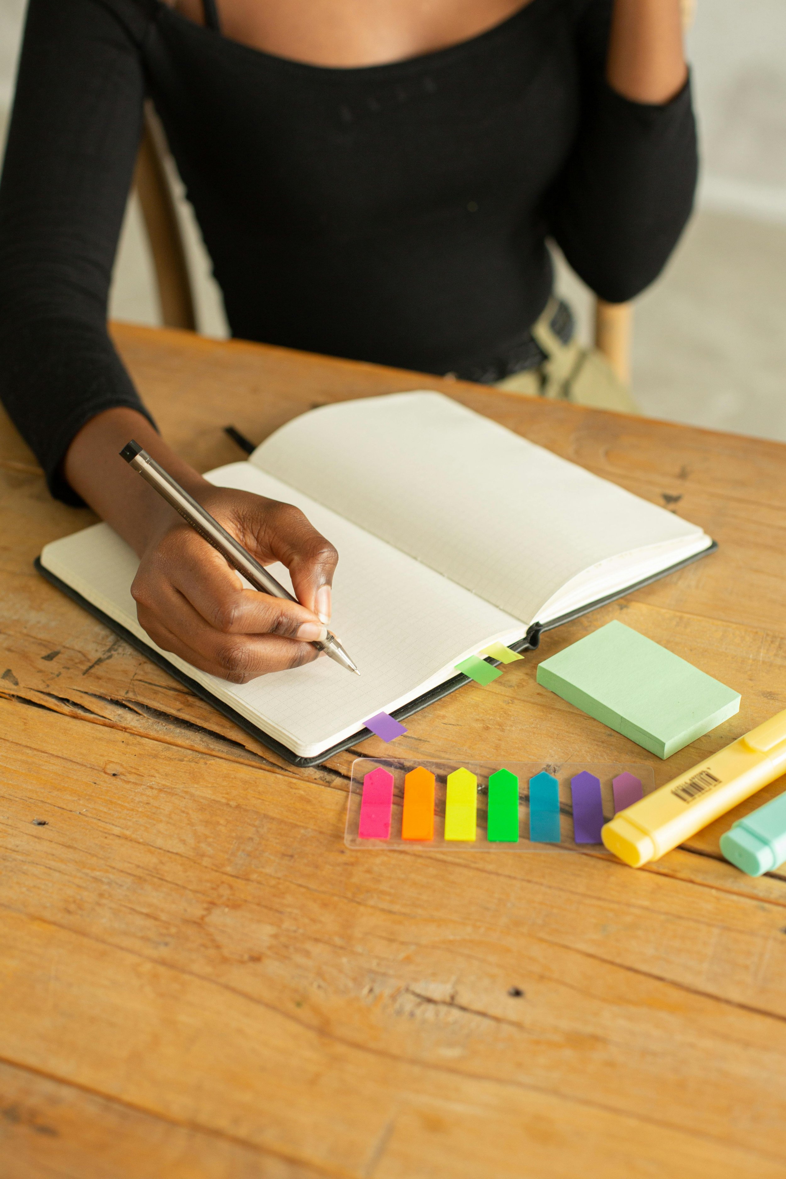 Person writing in notebook on wooden table surrounded by sticky notes and highlighters.