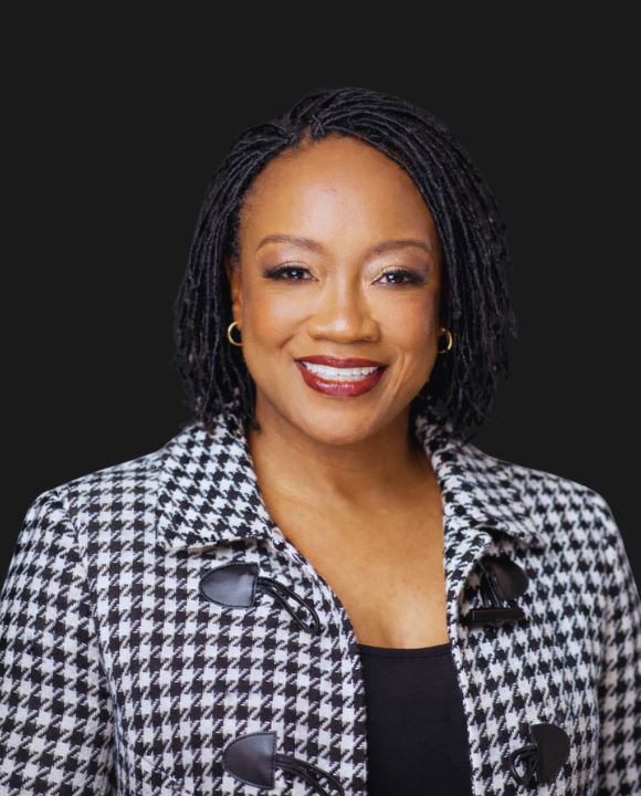 A woman with short dreadlocks wearing a black and white houndstooth jacket, gold hoop earrings, and a black top, smiling against a black background.