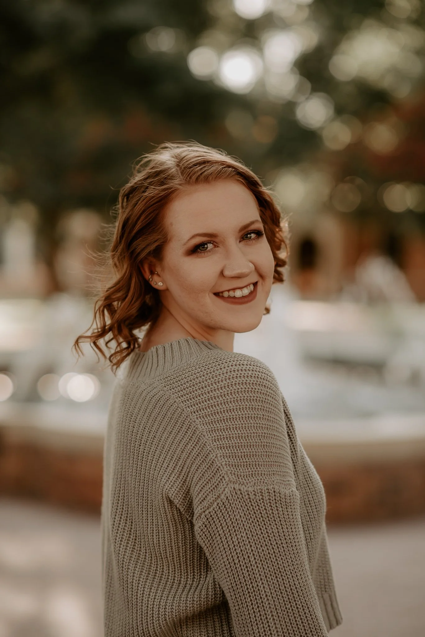 A young woman with short, wavy red hair and fair skin is smiling and looking at the camera outdoors during daytime. She is wearing a beige knit sweater, and there are blurred trees and boats in the background.