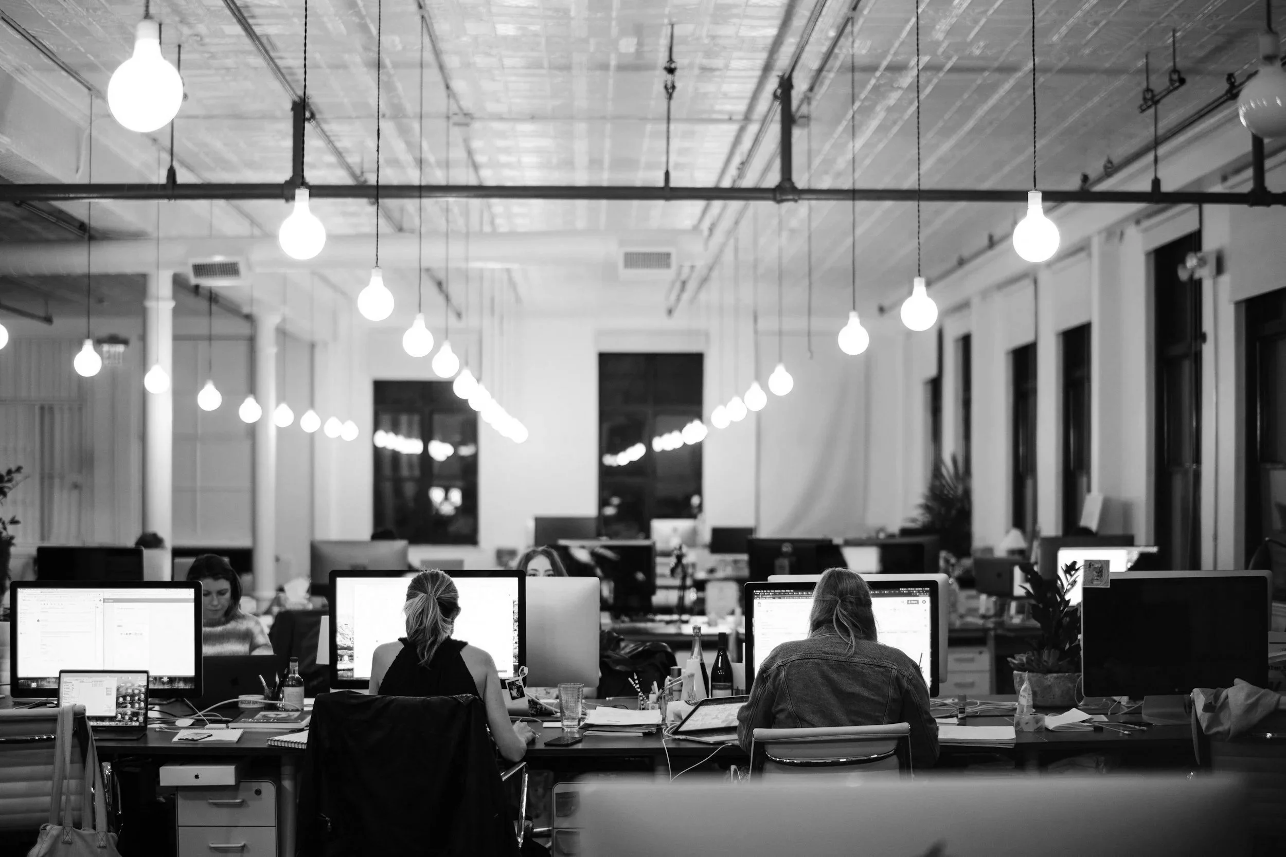 An office with multiple workers at desks working on computers. The office has high ceilings with hanging light bulbs and large windows. The image is in black and white.