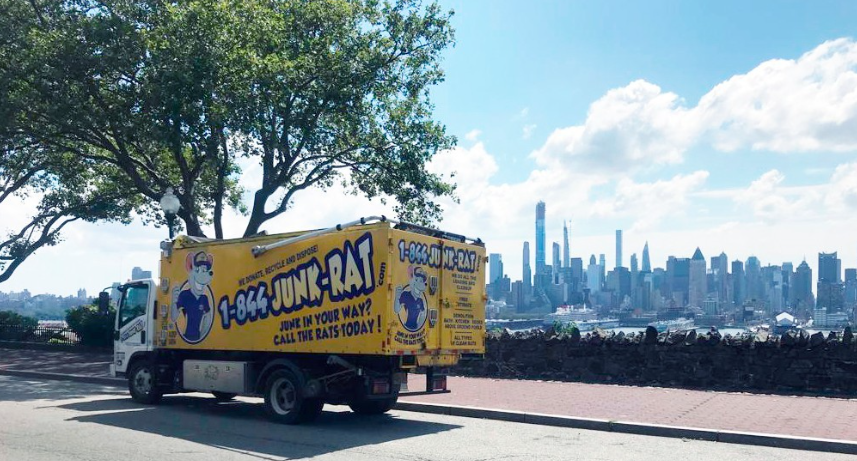 A yellow Junk-Rat truck parked on a street with a city skyline in the background, including tall skyscrapers and blue sky with clouds.