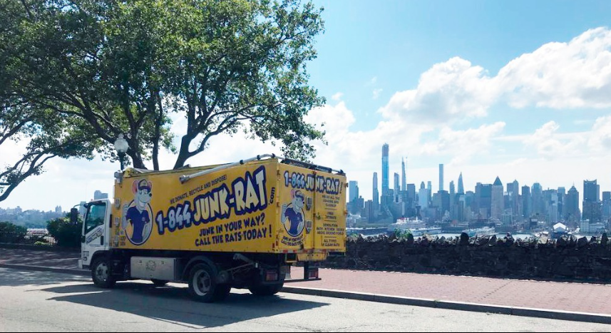 A yellow junk removal truck with the logo "Junk-Rat" parked on a city street, with a skyline of skyscrapers in the background under partly cloudy skies.