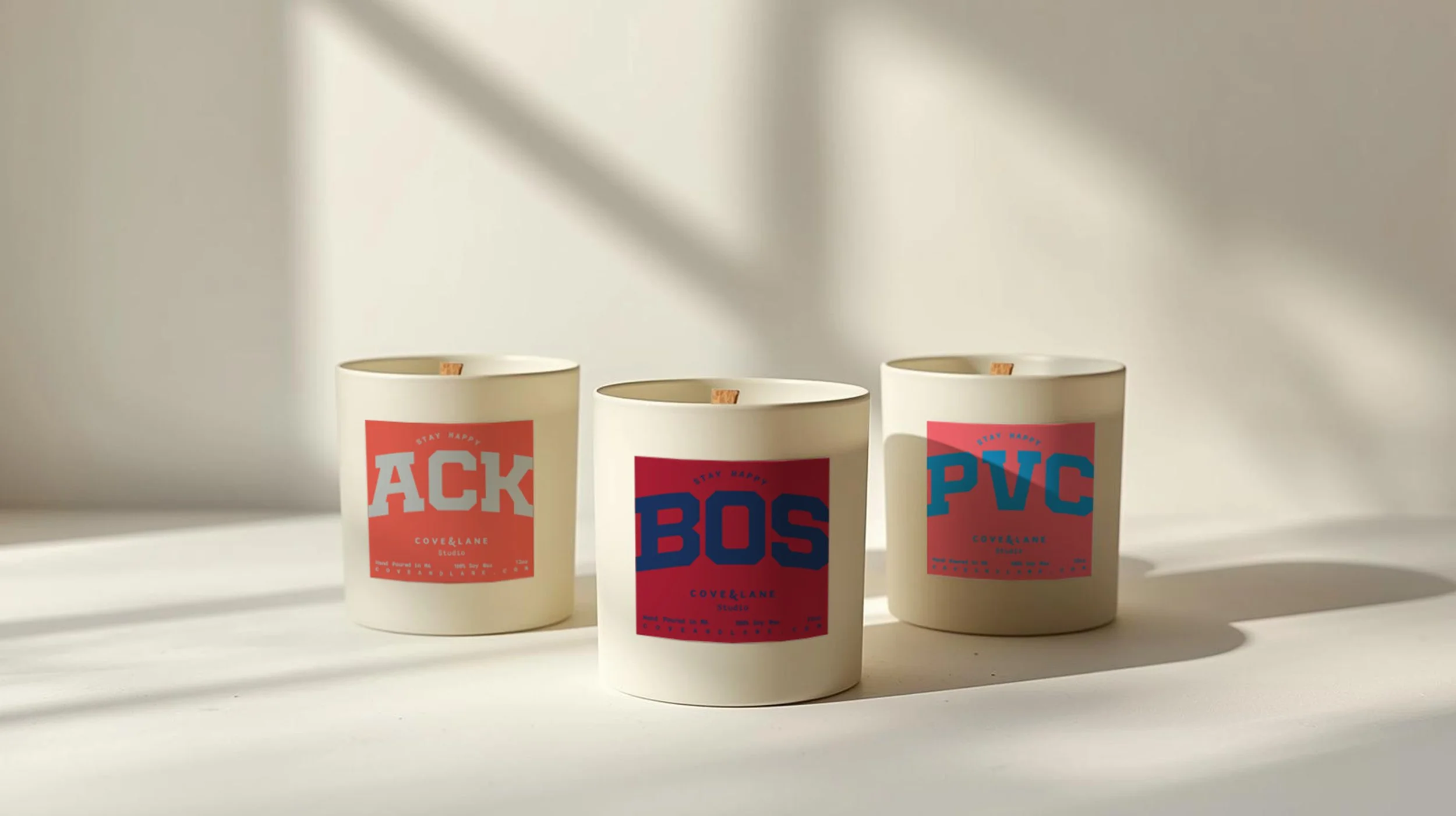 Three 12oz Happy Places Collection white candles with red and blue labels reading "ACK," "BOS," and "PVC," placed on a white surface in natural light.