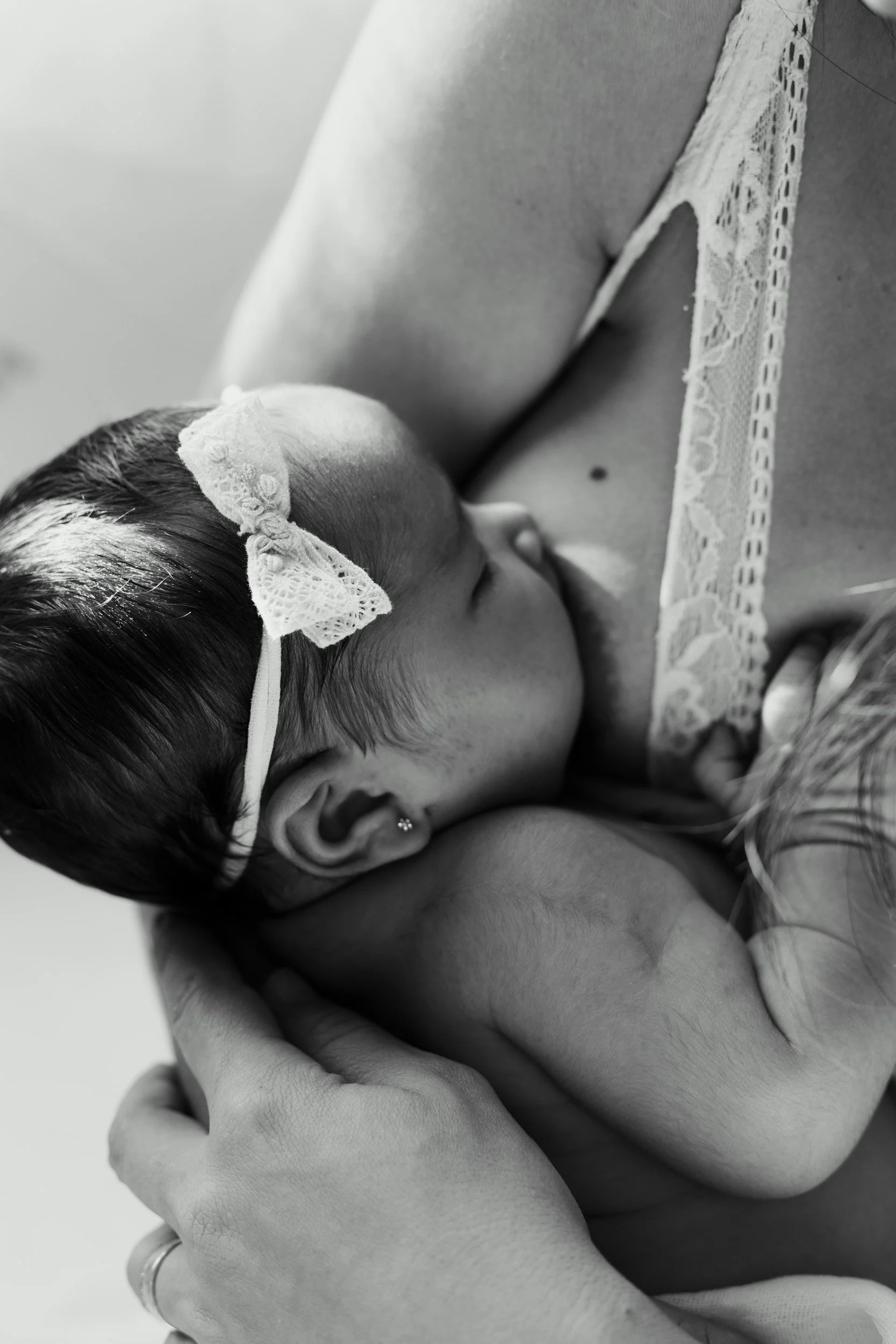 A black and white photo of a sleeping baby girl with a lace bow on her head, resting on a woman’s shoulder.