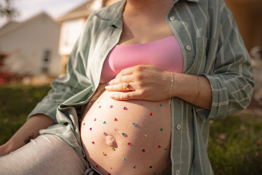 Close-up of a pregnant woman outdoors with a pink top and open green plaid shirt, decorated with colorful triangle stickers on her belly, holding her belly with one hand.
